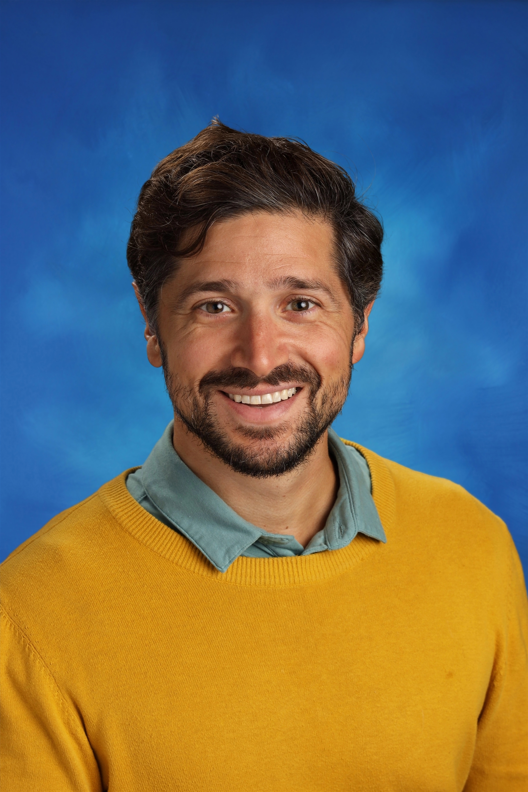 Anthony Aceti wears a yellow sweater on a blue background while smiling warmly.