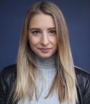 Stephanie appears on a dark grey background. Her long blond hair reaches down over a light grey turtleneck and black leather jacket. Stephanie looks at the camera with a gentle smile, hinting at her playful energy.