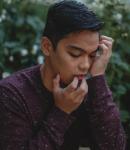 A side profile of Miggy, who crouches amidst bushes and white flowers that recede blurrily into the background. His fingers gently crawl up from his long-sleeved maroon shirt, over his chin and lips, and toward his black, wavy hair. The brown skin of his cheek is caressed by his palm as he looks down with eyes closed in contemplation.